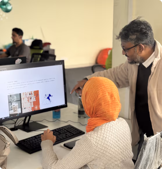A man wearing glasses and a beige jacket points at a computer screen while a woman in an orange headscarf types on the keyboard in an office setting.