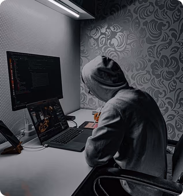 Person in a hoodie working at a desk with a laptop and an external monitor displaying code in a dimly lit room.