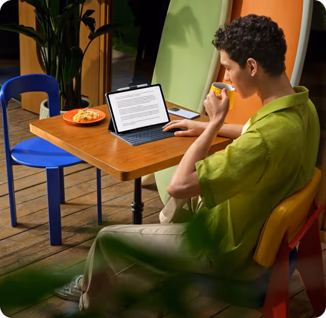 A young man in a green shirt sitting at a wooden table, working on a tablet with a keyboard while drinking from a yellow cup.