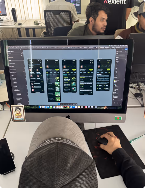 Person wearing a cap working on a desktop computer displaying a mobile app design interface while others work in the background.
