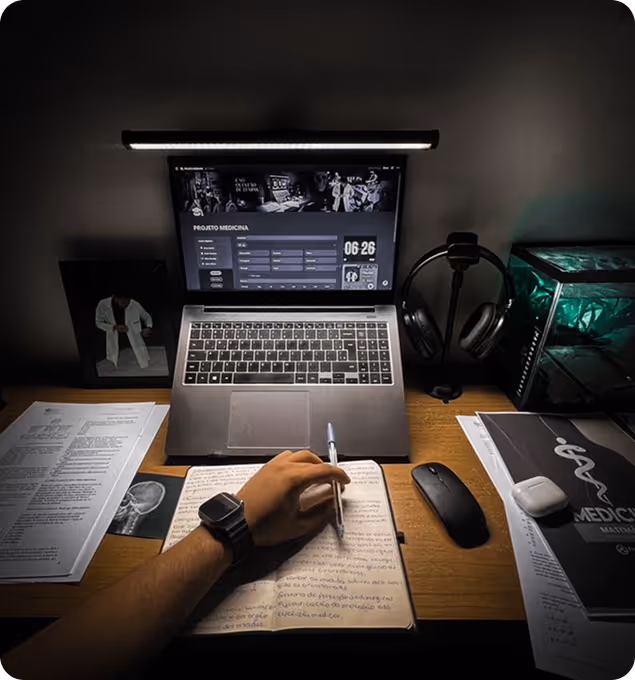 Person writing notes in a notebook at a desk with a laptop displaying a medical project, surrounded by documents, a mouse, headphones, and a small decorative item.